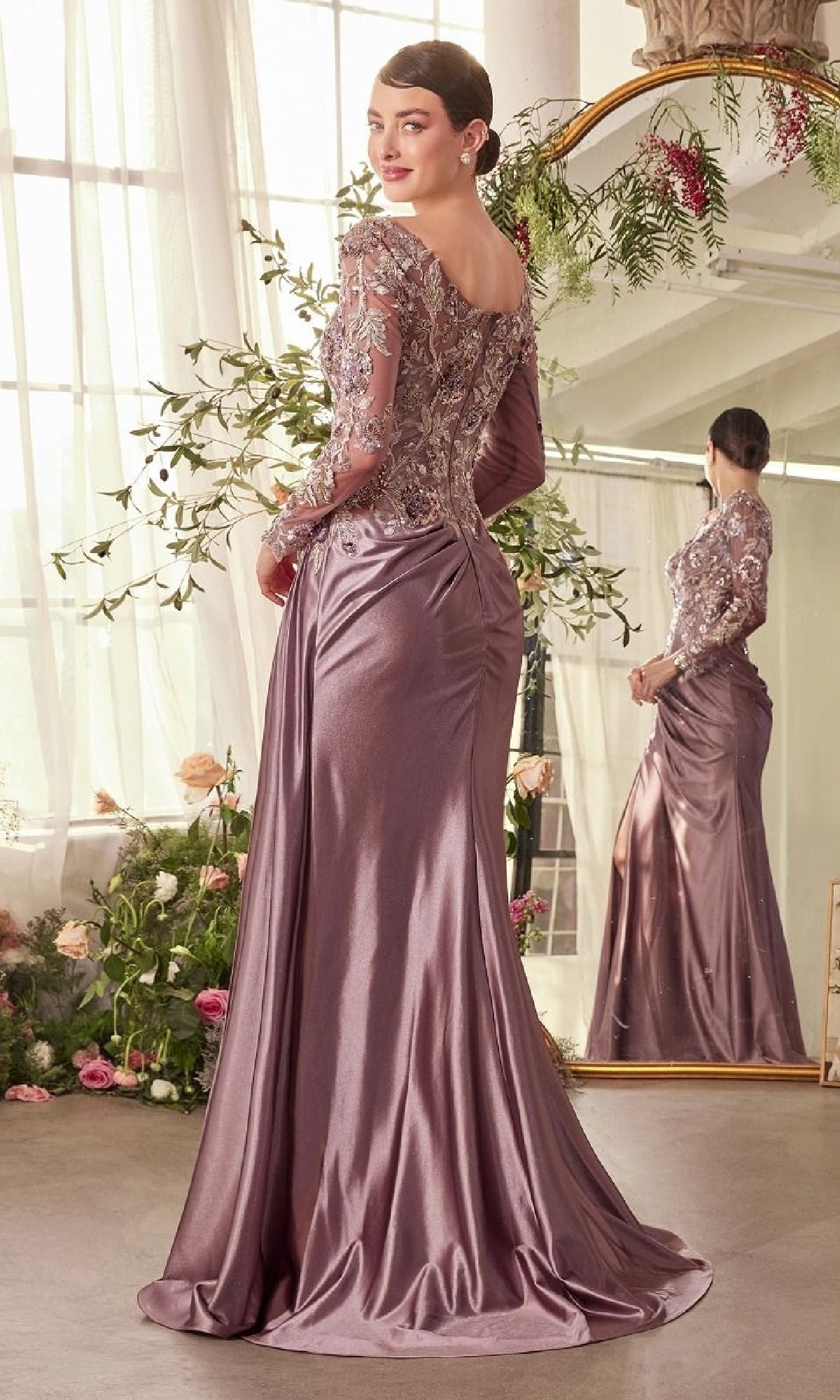 A woman wearing the Andrea and Leo A1403 long formal dress, featuring a mauve satin ruched skirt and sheer embellished sleeves, stands in front of a mirror in a floral-decorated room.